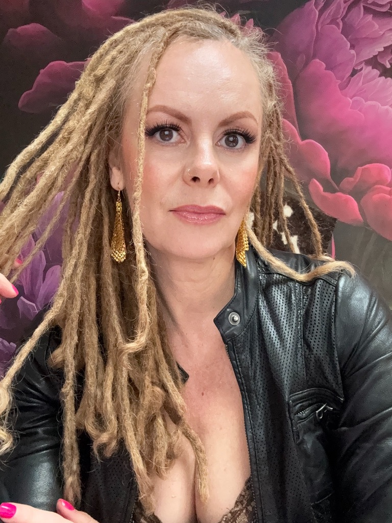 Marta Kagan in a black leather jacket with gold earrings, in front of large magenta flowers
